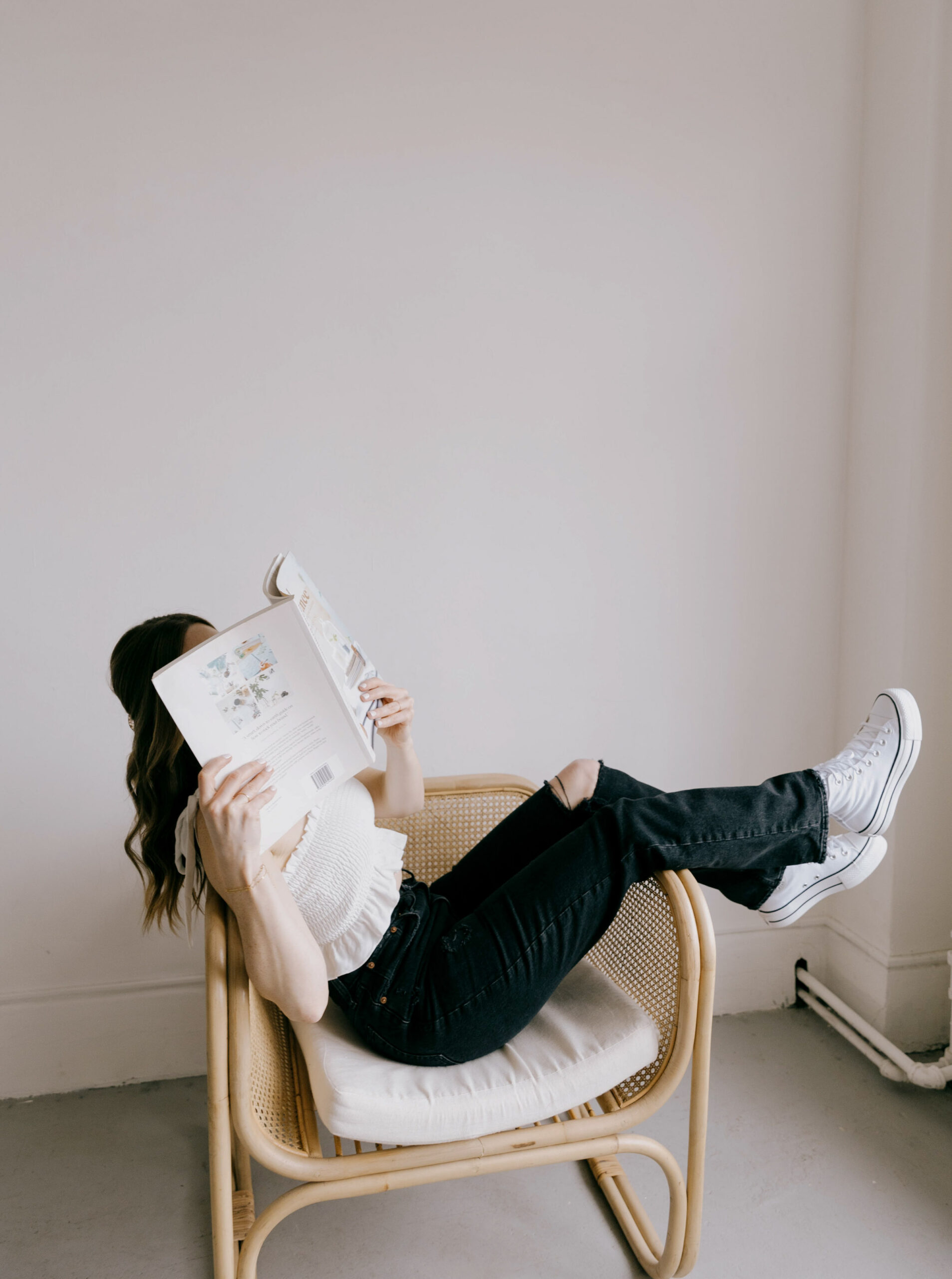 A woman lounging in a rattan chair with her legs up, reading a book that covers her face. She’s wearing a white smocked top, black ripped jeans, and white platform sneakers, set against a minimalist white wall in a relaxed, modern space.