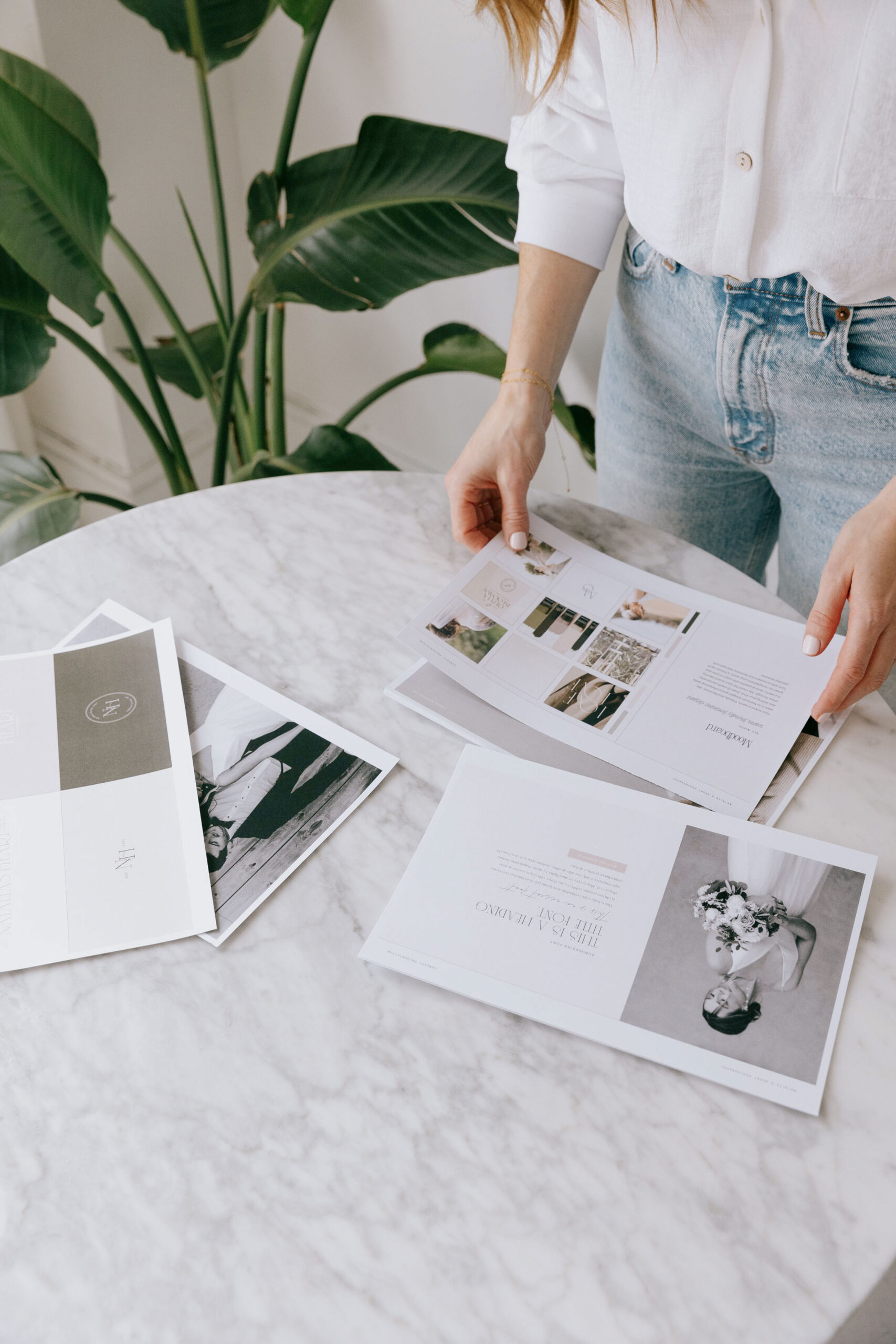 A woman in a white blouse and light denim jeans arranging printed brand design mockups on a marble table, surrounded by tropical green plants. 